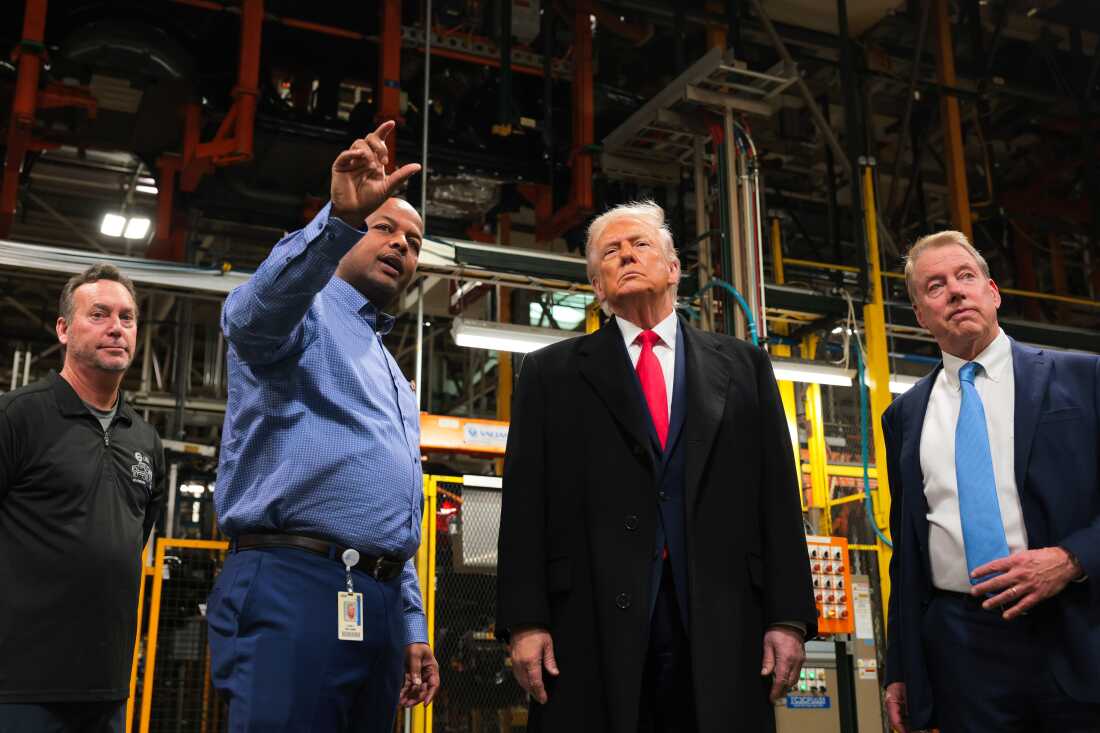 President Trump stands next to employees at the Ford River Rouge plant, as well as Ford Motor's executive chair, Bill Ford Jr., who's standing on the right side of the frame. One employee, on the left side of the frame, has one arm raised in a gesture. Metal factory equipment stands in the background.