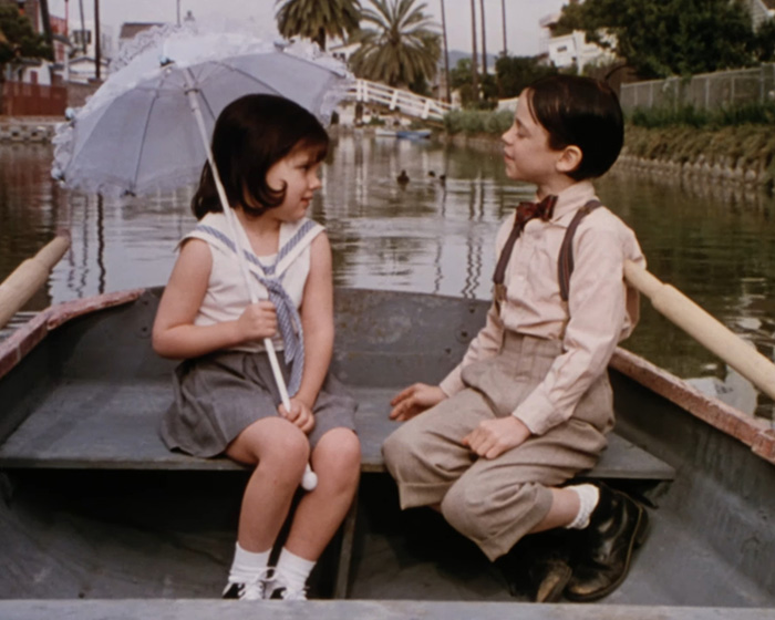 Two children dressed in vintage clothing sitting in a boat near a canal, reflecting Little Rascals star living off the grid.