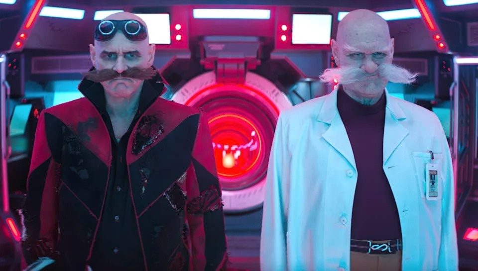 Two characters in futuristic costumes, one in a black suit with goggles, the other in a white lab coat, stand in a high-tech setting