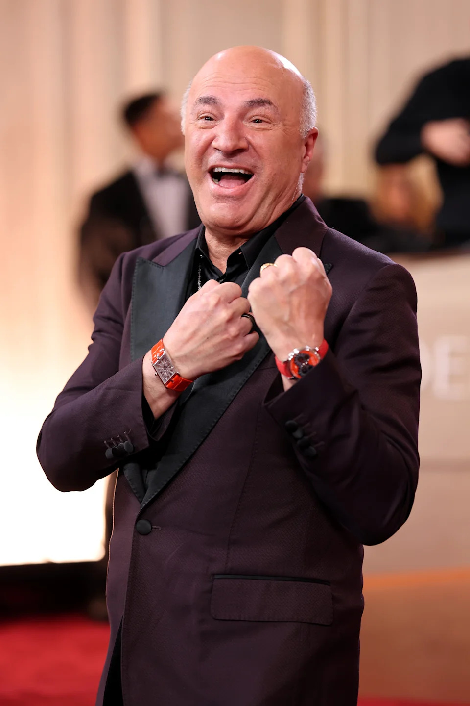 Person in formal suit on red carpet, smiling broadly with fists raised, wearing a watch and bracelet