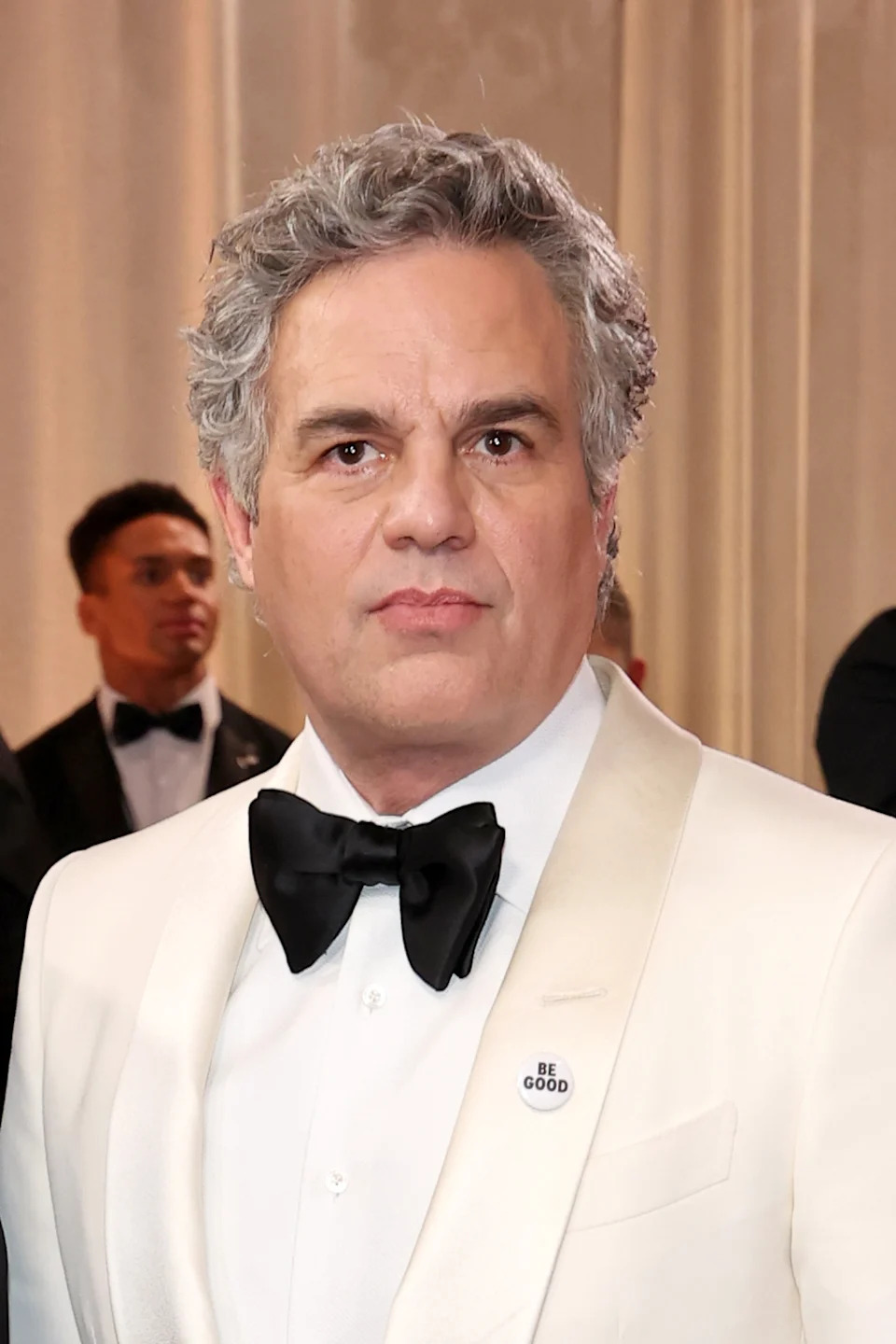 A celebrity in a white tuxedo with a black bow tie on a formal event backdrop. A "Be Good" pin is visible on their lapel