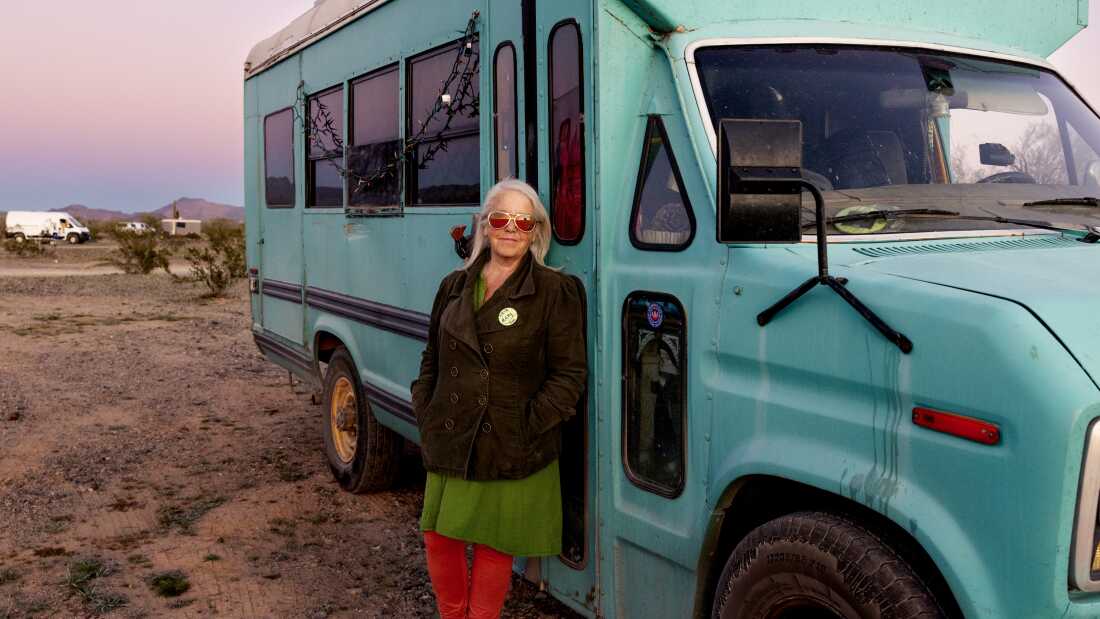 Mary Freuer, who has shoulder length gray hair and is wearing sunglasses, stands outside of her teal converted short school bus, also known as a skoolie, as the sun sets.