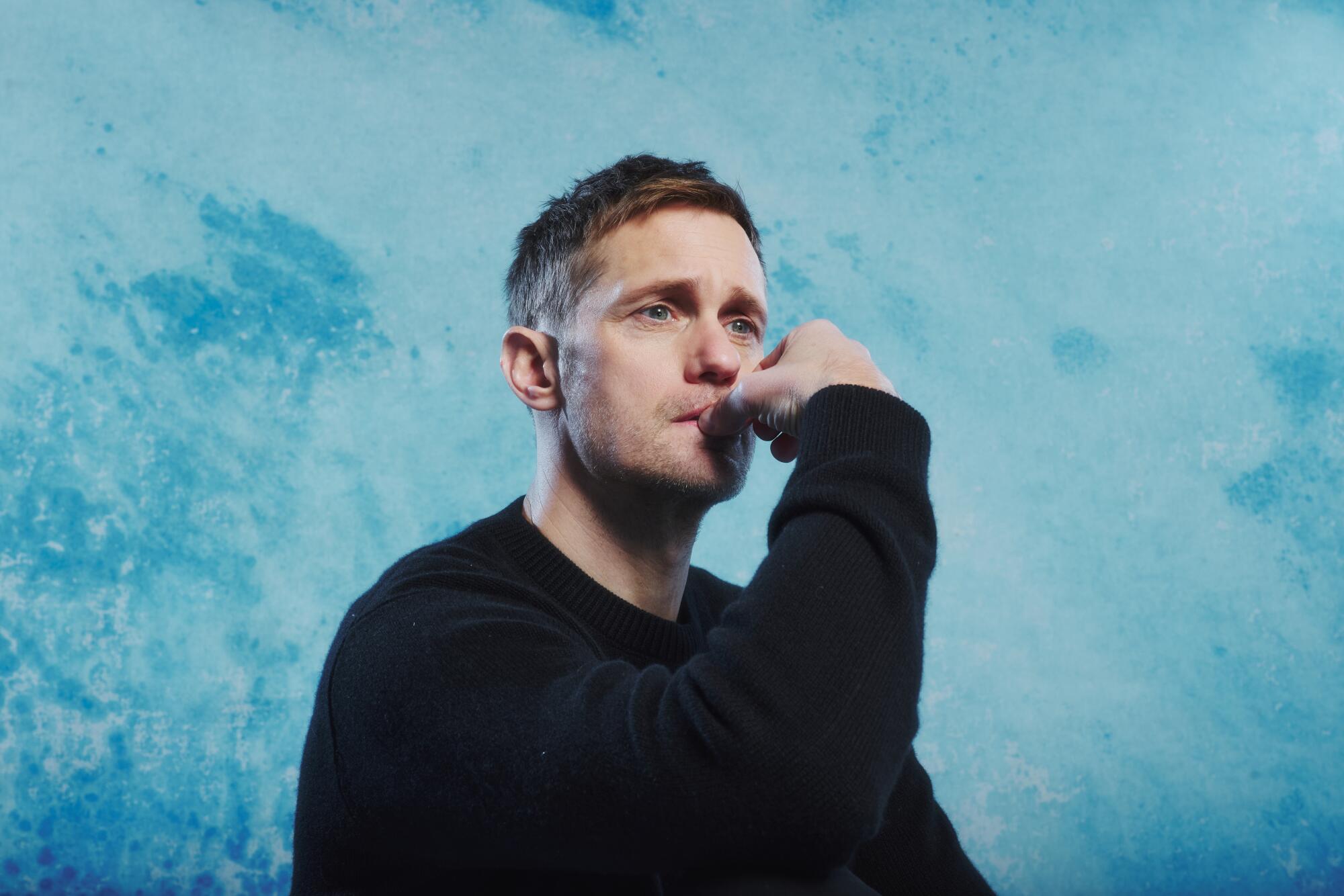 A man in a dark top touches his lips with his hand.