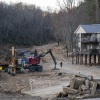 A precarious-looking wooden apartment complex is shown on stilts, on ground where a river flooded and receded. Big construction equipment is shown in front of the building.