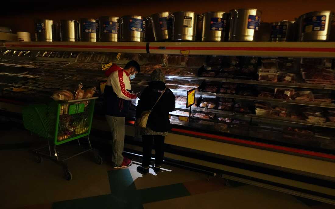 In this Tuesday, Feb. 16, 2021 photo, customers use the light from a cell phone to look in the meat section of a grocery store that was without power, in Dallas. Brazos Electric Power Cooperative Inc., the oldest and biggest generation and transmission power cooperative in Texas has filed for Chapter 11 bankruptcy protection following last month’s winter storm that left millions without power. (AP Photo/LM Otero, File)