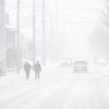 Two individuals walk along a street; they, the vehicles behind them, and the building next to them are blurred by snowfall.
