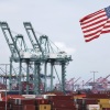 Large shipping containers are stacked at the Port of Los Angeles. Cranes for moving them stand in the background, and a U.S. flag flies in the foreground.