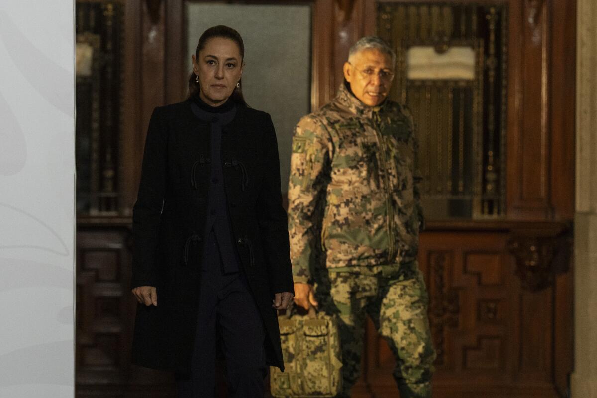 A woman with dark hair, in dark clothes, standing near a man with gray hair, in fatigues, holding a briefcase