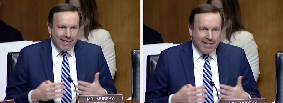 A man in a suit and tie is speaking animatedly during a meeting, seated at a table with a nameplate visible