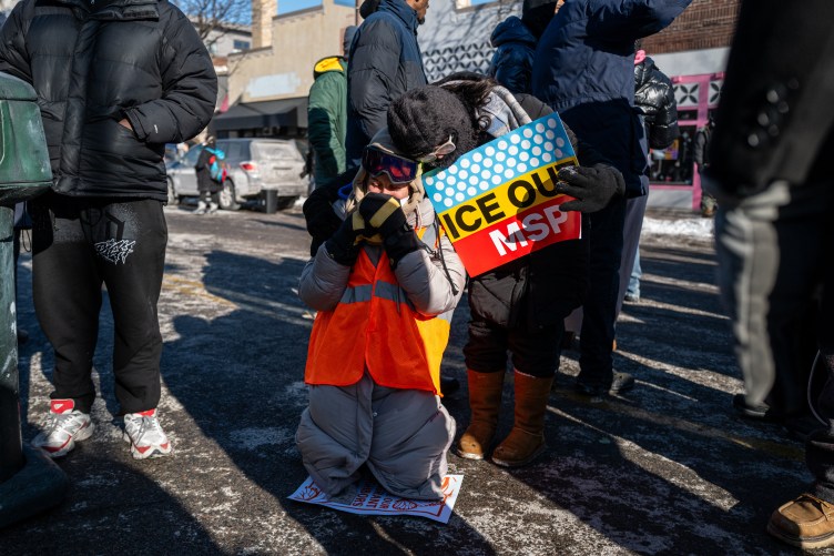People wearing winter clothing stand in a street. One woman, wearing an orange vest and goggles, is kneeling on the ground with her hands clasped toward her face. Another woman leans down to put her arm around the kneeling woman. She is holding a sign that says “Ice Out MSP.”