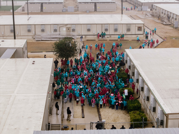 A dense crowd of hundreds of people wearing raincoats and hoods is seen from an aerial perspective. Many of them are holding signs.