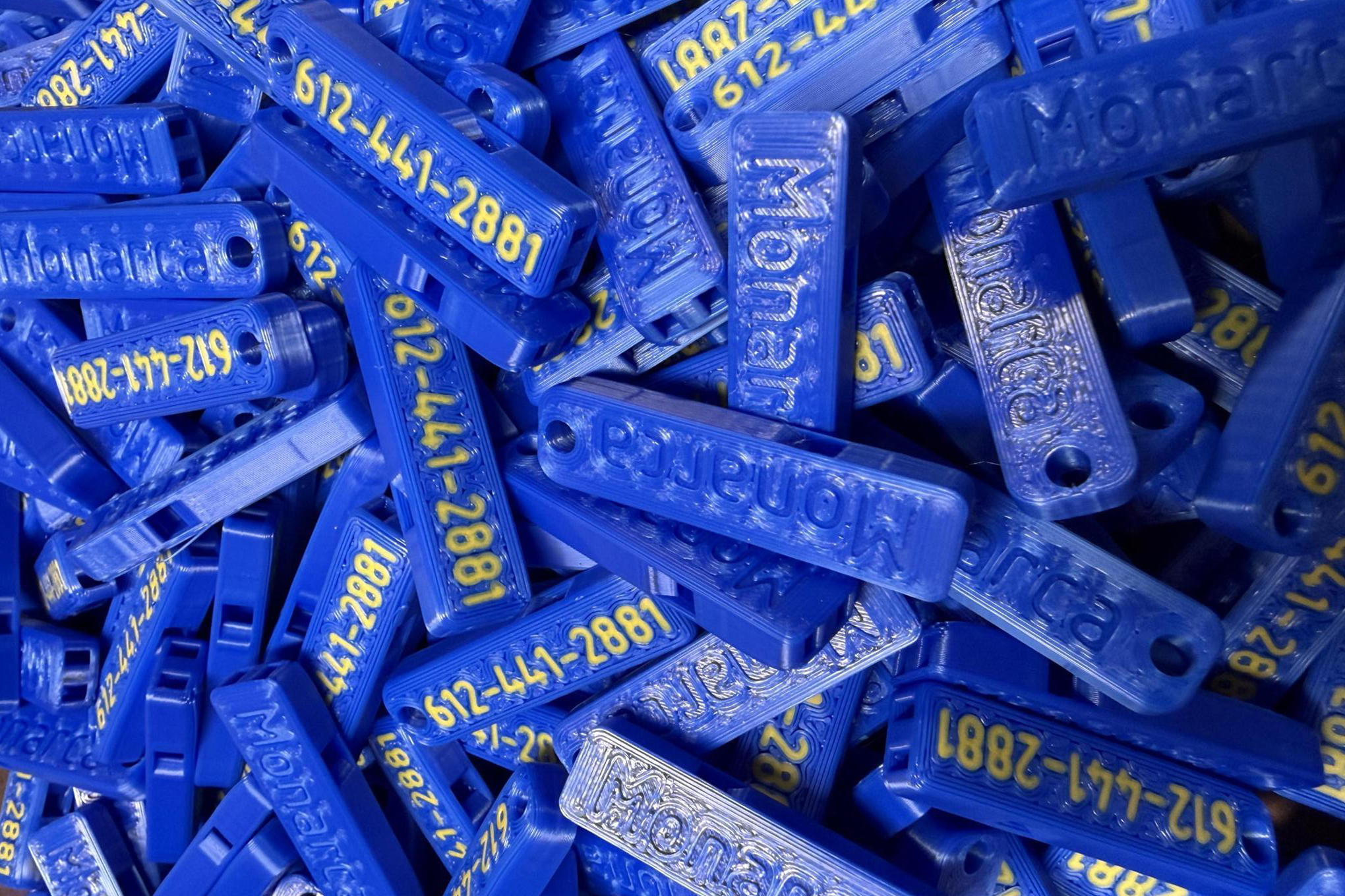 A pile of blue 3D-printed whistles in every orientation, with the phone number 612-441-2881 pirinted on one side and “Monarca” on the other.