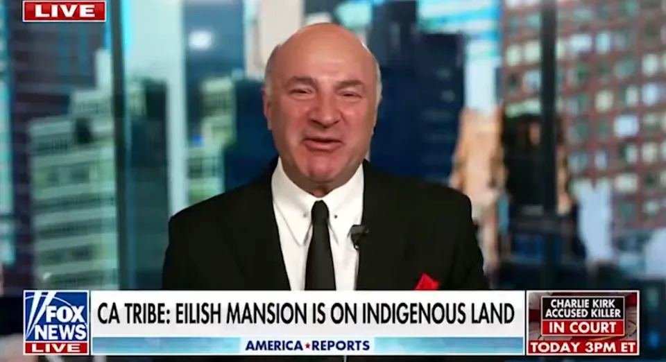 Man in a suit on a news broadcast with headline about Billie Eilish's mansion on indigenous land; ticker mentions Charlie Kirk in court