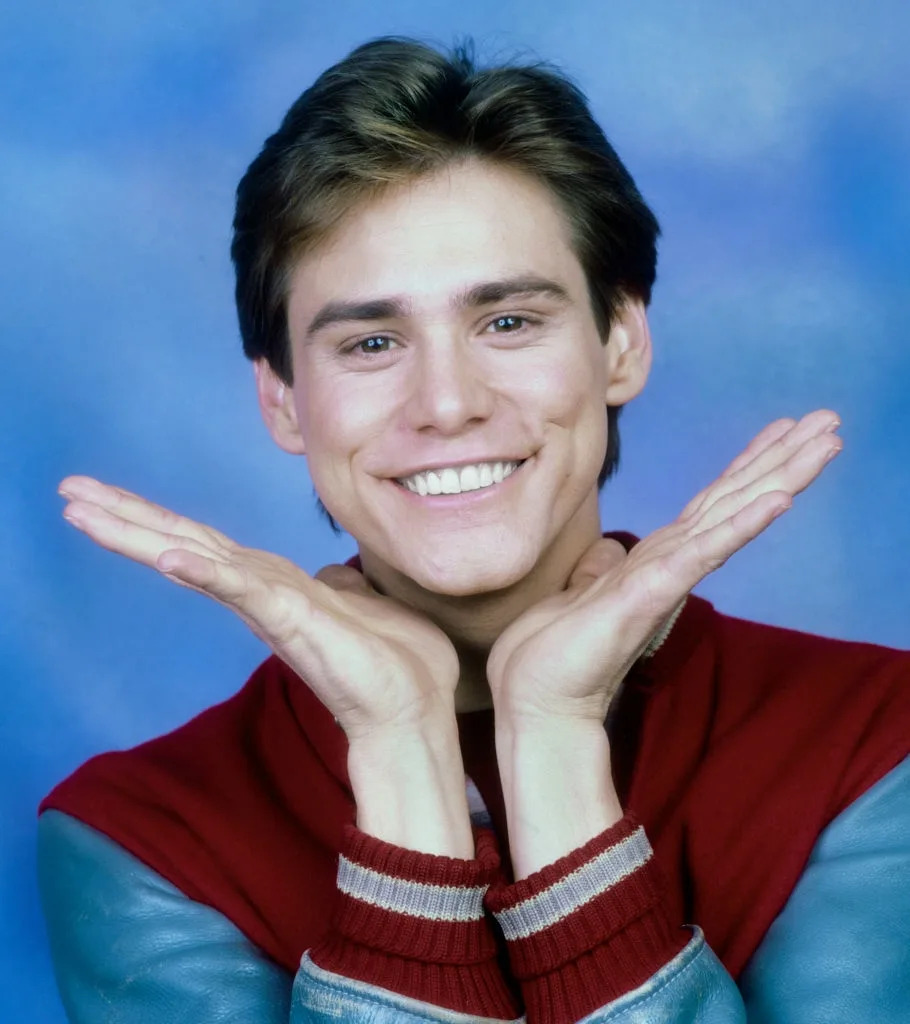 Person posing playfully with hands framing their face against a blue background, wearing a casual jacket