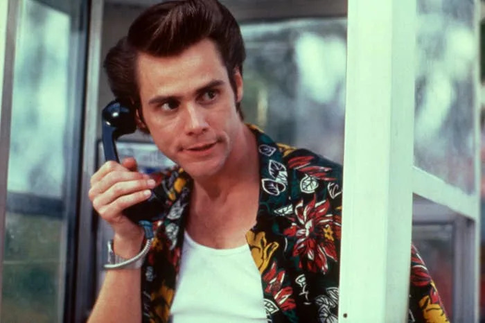 A person in a floral shirt and white undershirt holds a phone inside a phone booth, appearing to listen attentively