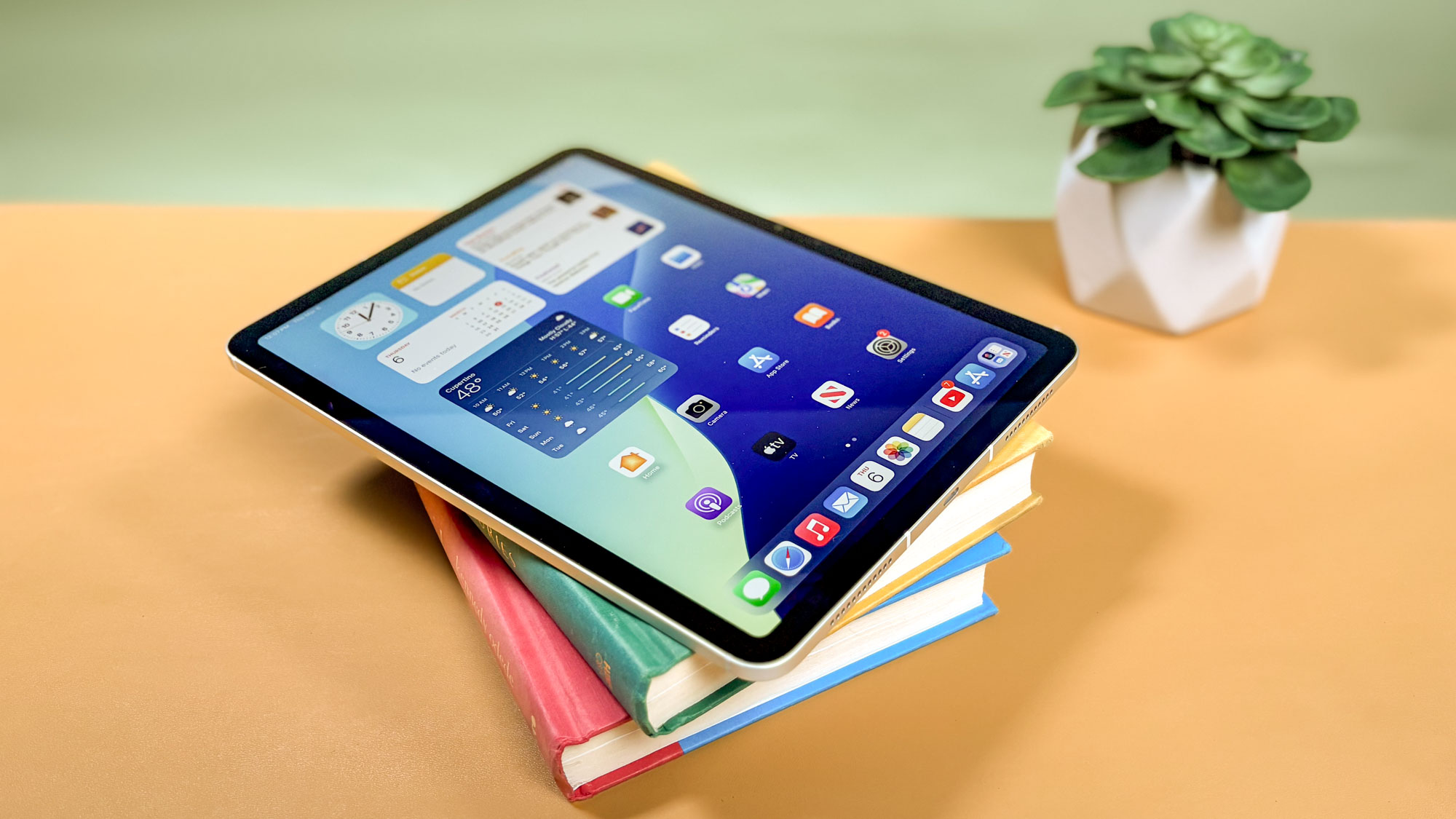 iPad Air M3 on top of a pile of books stacked at an angle on a yellow office table next to a houseplant