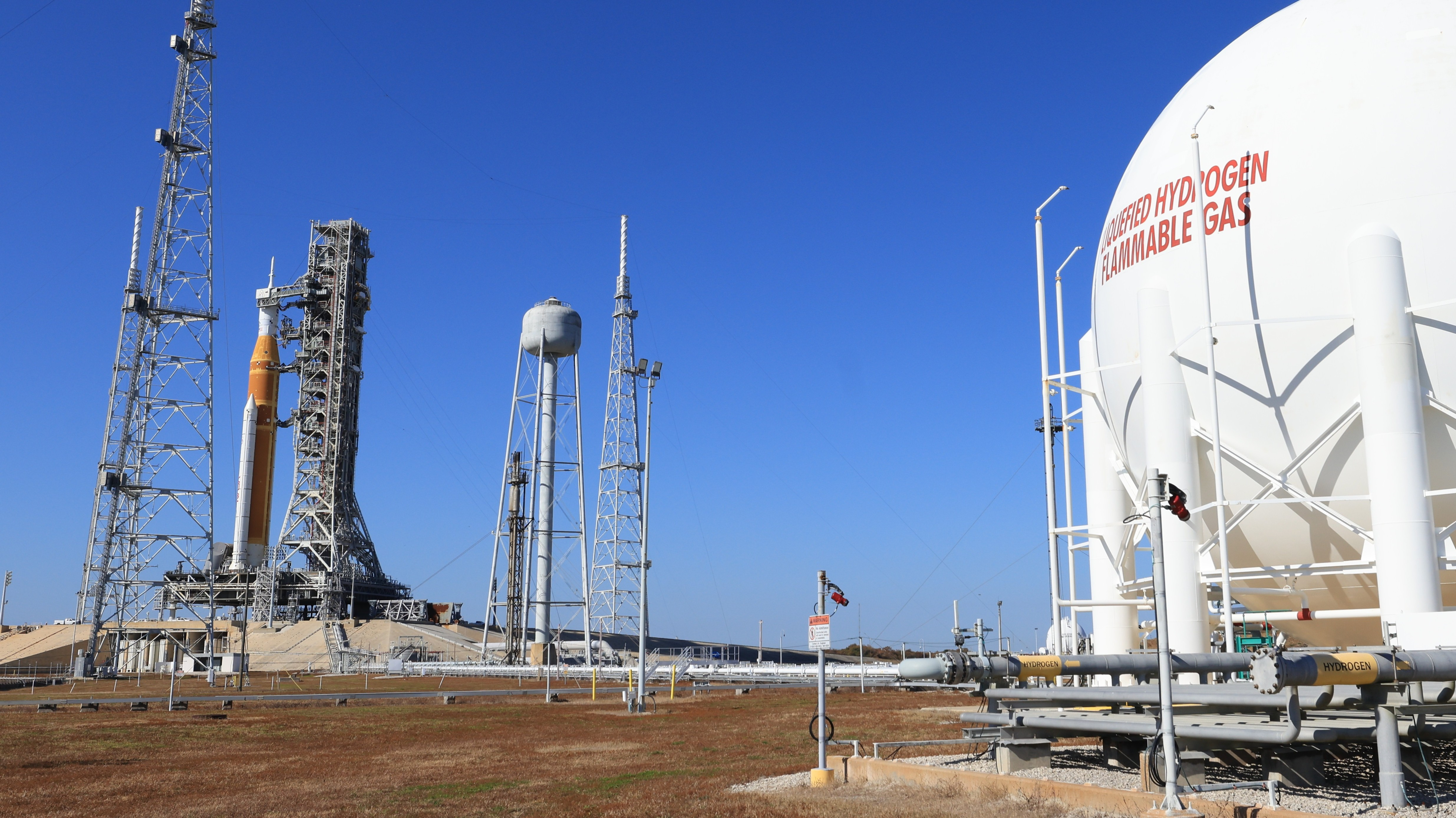 a tall orange rocket stands in the distance on the left, with a large white fuel tank in the foreground on the right.