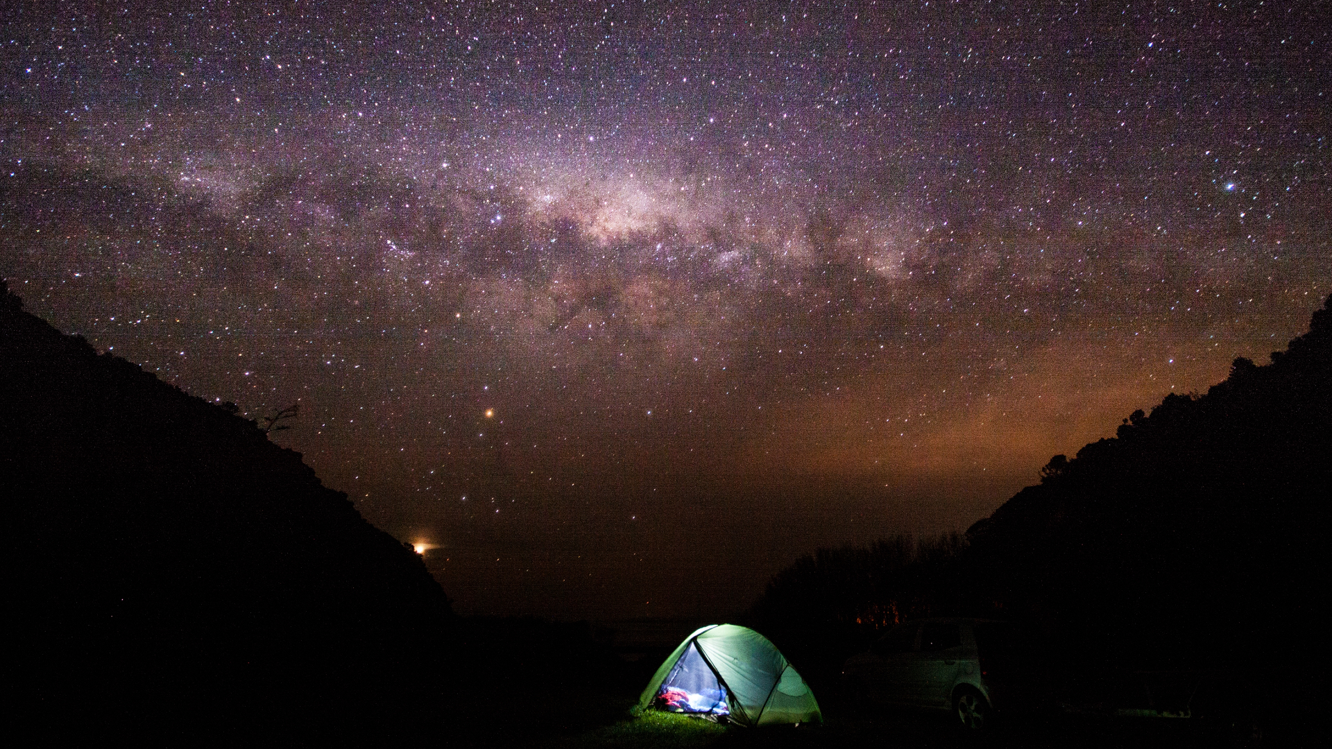 A green tent is illuminated by a flashlight next to the silhouette of two sloping hills with an orange starry night sky and a purple and pink streak of the Milky Way galaxy glowing above