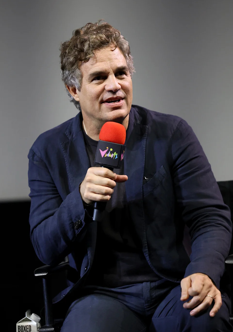A person in a dark suit speaks into a colorful microphone while seated at an event