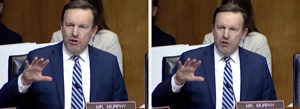 Man in a suit and tie speaks energetically, gesturing with one hand during a formal event. Nameplate reads "Mr. Murphy."