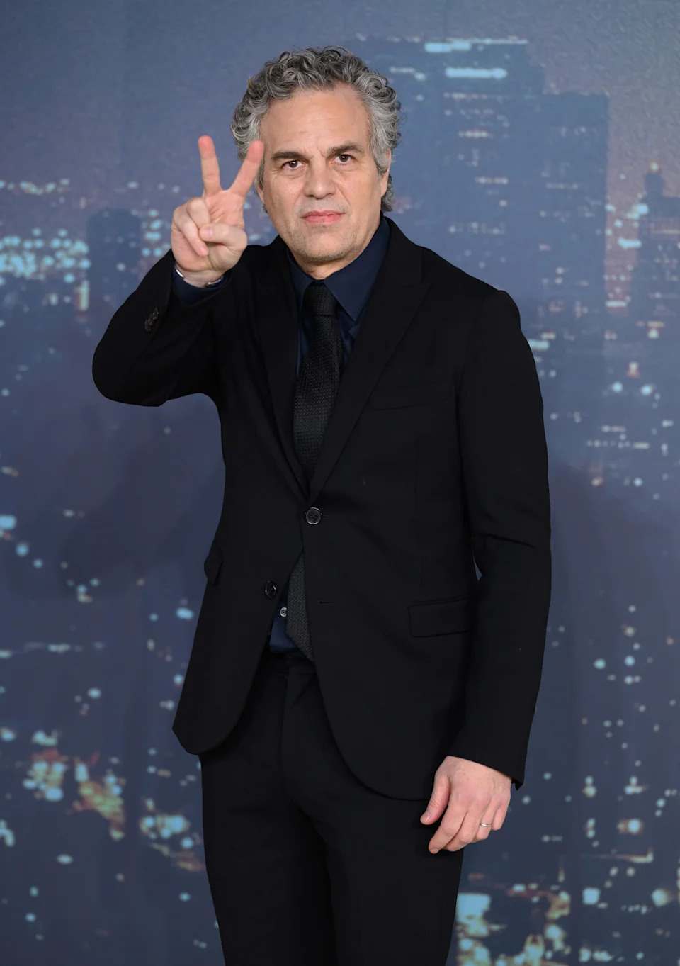 A man in a dark suit poses on a red carpet, holding a peace sign with his fingers against a cityscape backdrop