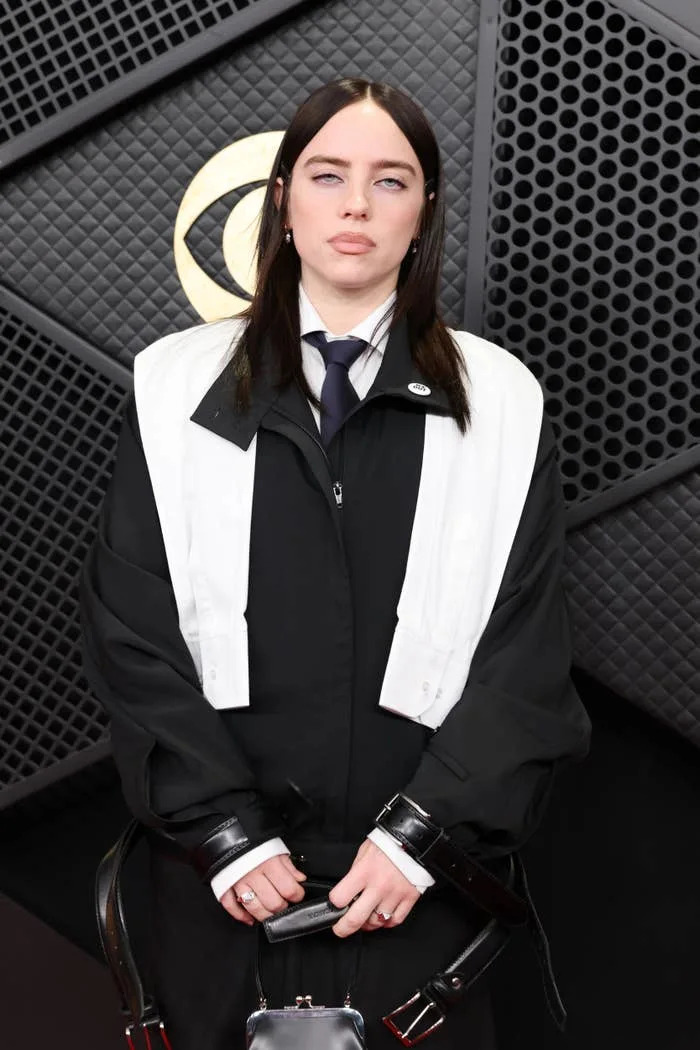 Person wearing a black coat with a white cape and tie at a celebrity event. Background features a textured pattern with a circular design