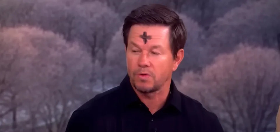 A man with an ash cross on his forehead appears on a talk show, wearing a dark shirt. Trees are visible in the background