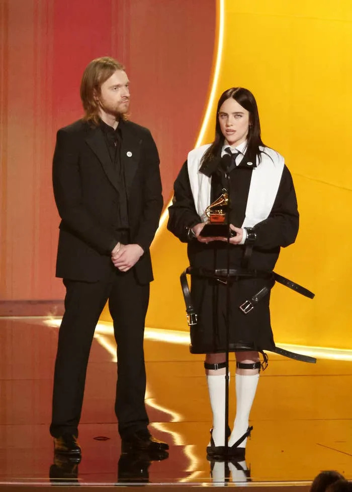Two people on stage at an awards event; the one on the right holds a trophy and wears a unique black and white outfit with straps and shorts