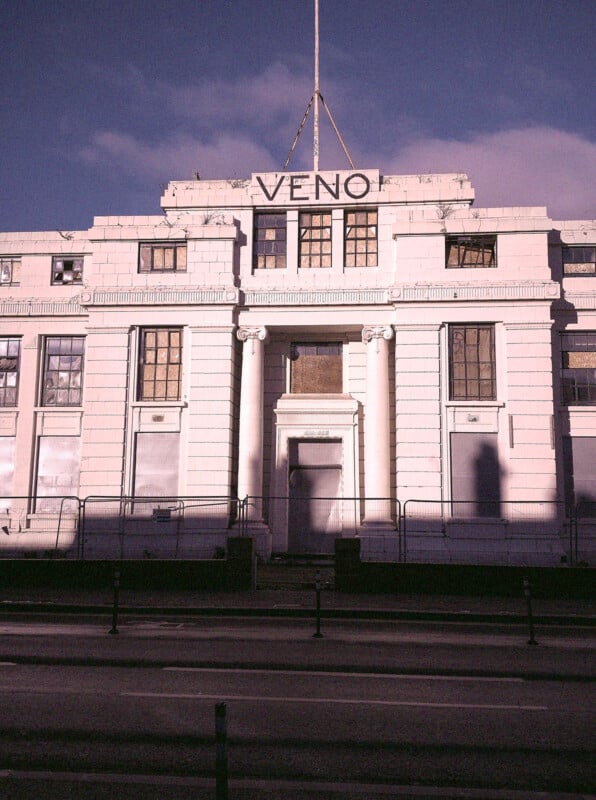 A white, neoclassical-style building with tall columns and boarded-up windows. The word “VENO” is displayed in large letters on the roof. The building appears abandoned and is surrounded by barriers.