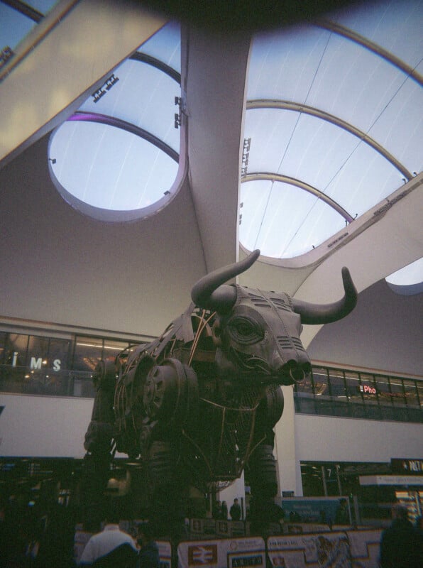 A large mechanical bull sculpture stands inside a modern building with circular skylights, surrounded by shops and people. The bull is dark with visible gears and wires, towering under the curved glass roof.