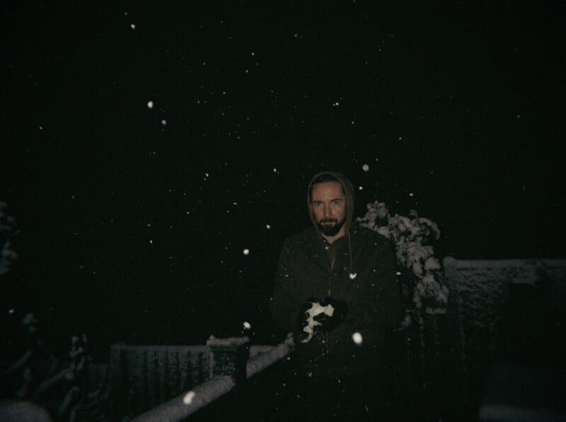 A man in a hoodie stands outside at night holding snow, surrounded by falling snowflakes and dimly lit by a soft light, with a snowy fence and plants in the background.