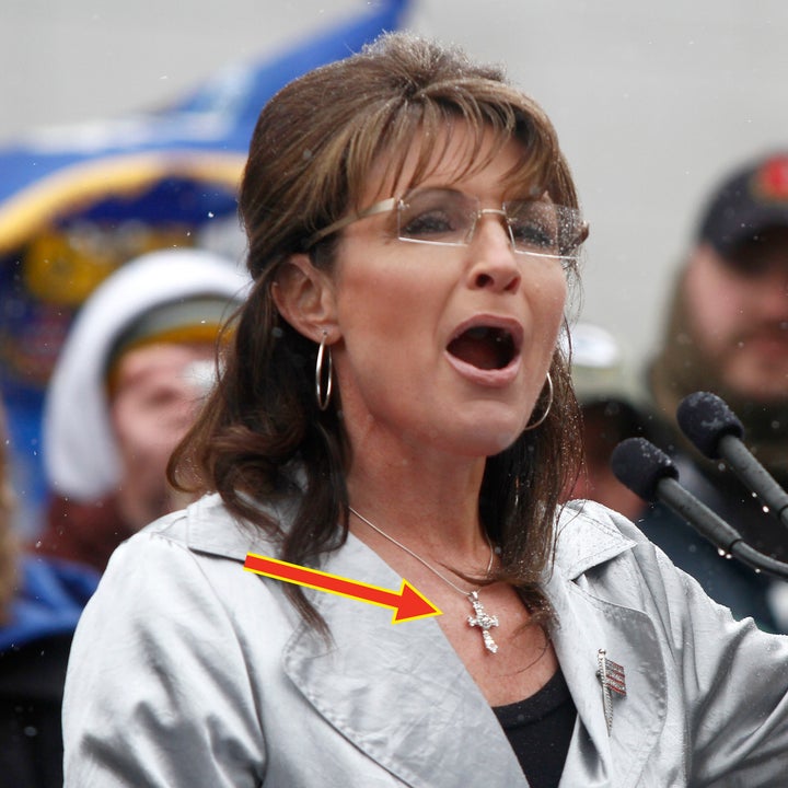 A woman speaks passionately at an outdoor event, wearing a shiny blazer and glasses, gesturing with her hand for emphasis