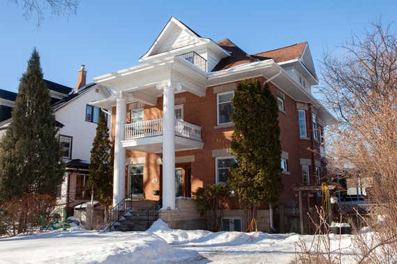 A red brick house with a white two-story pillared porch stands behind a snow-covered yard under a clear blue sky.