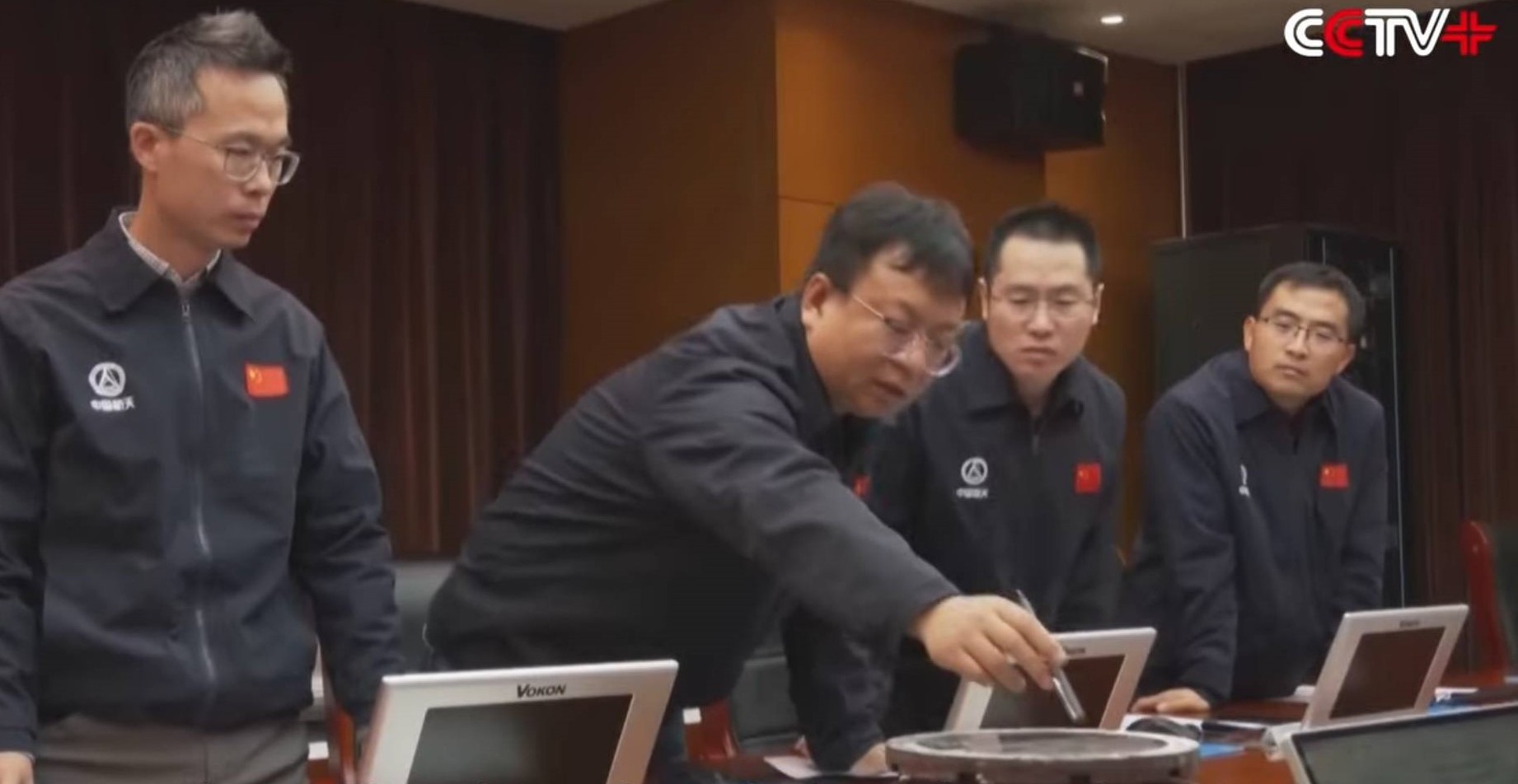 four men in black jackets point to a round pane of glass on a desk