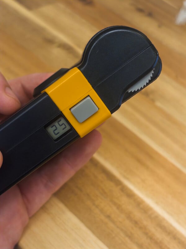 A close-up of a hand holding a black and yellow digital caliper or measuring tool, displaying "25" on a small digital screen, over a wooden surface background.