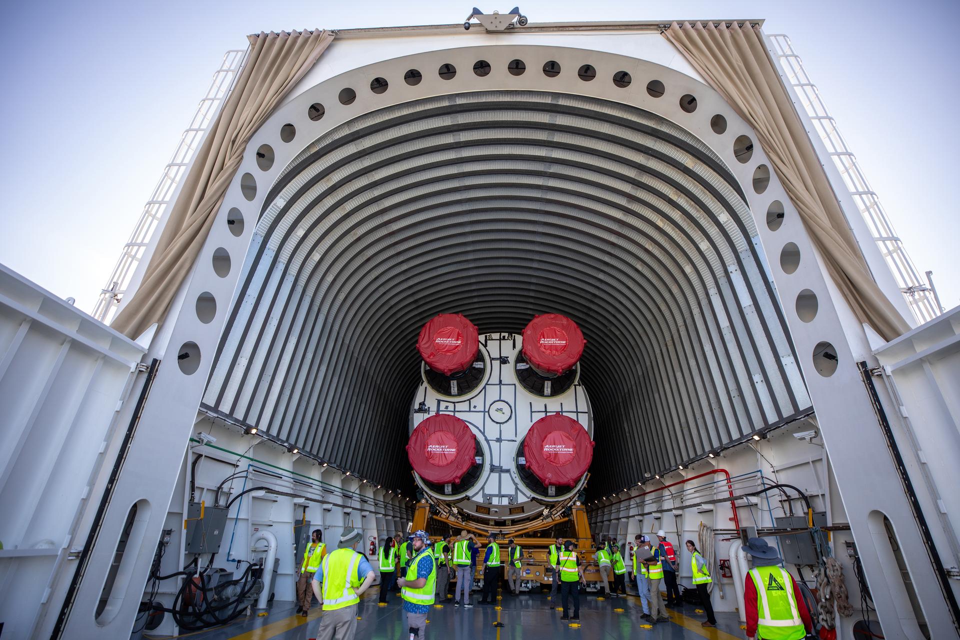 A team of workers in bright yellow vests surrounds a large rocket core stage, which is inside the cargo area of NASA's barge Pegasus.