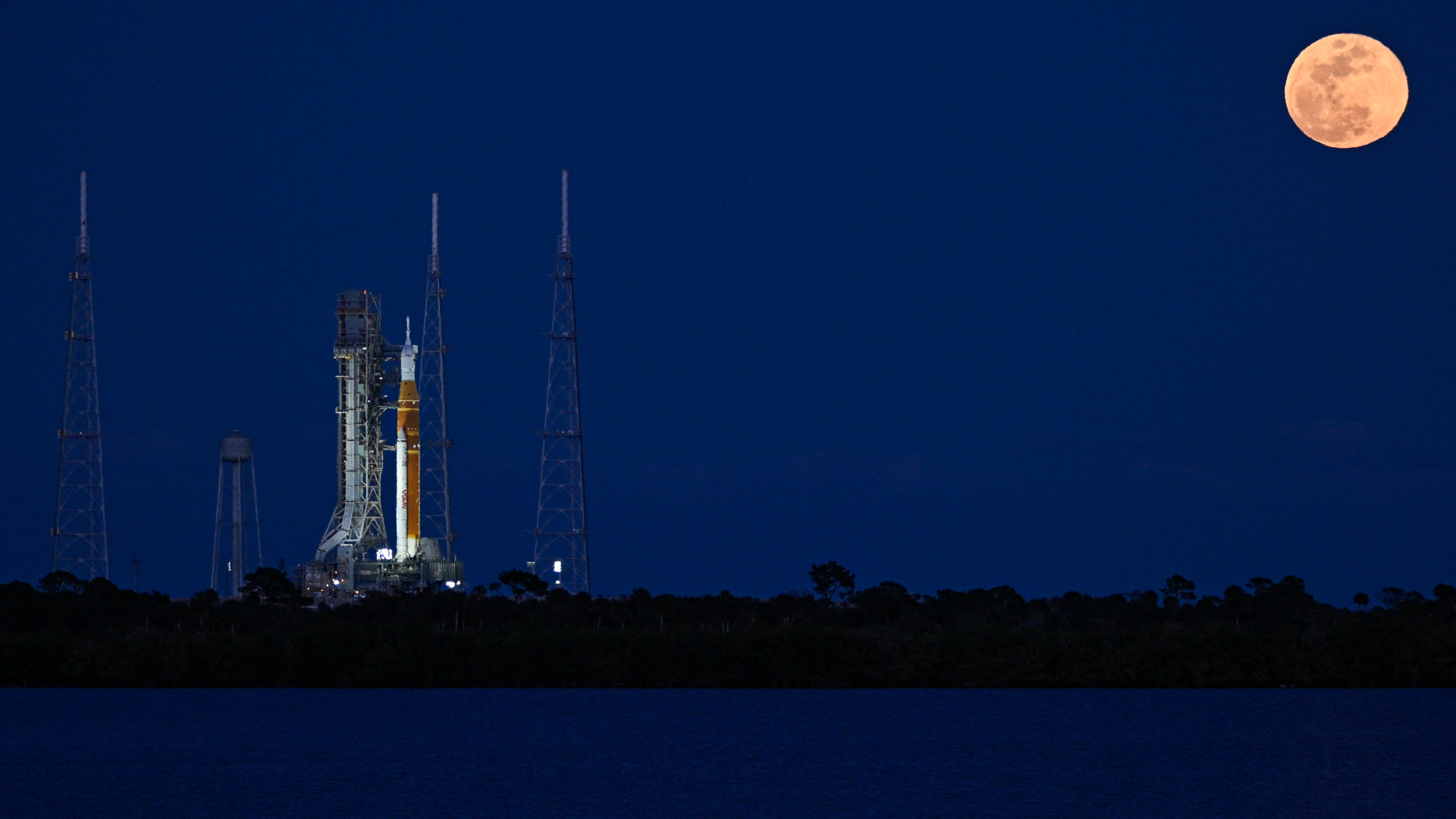 A full moon is pictured shining in a dark blue sky as a large rocket stands illuminated on a launch pad to the lower left.