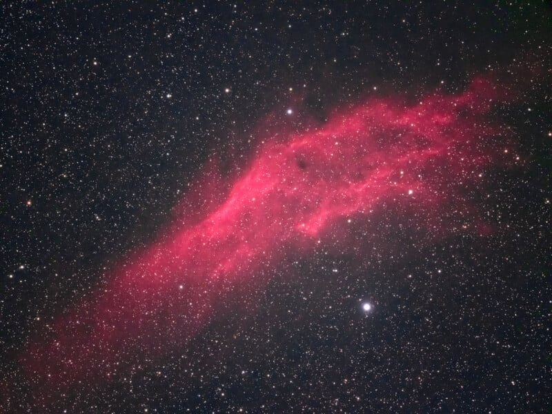 A bright reddish-pink nebula stretches diagonally across a star-filled black sky, with scattered stars of varying brightness surrounding the glowing cloud of gas and dust.