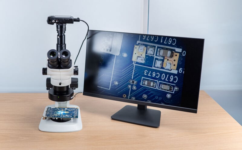 A digital microscope examines a circuit board on a desk; its camera is connected to a monitor displaying a magnified image of the circuit board’s electronic components.
