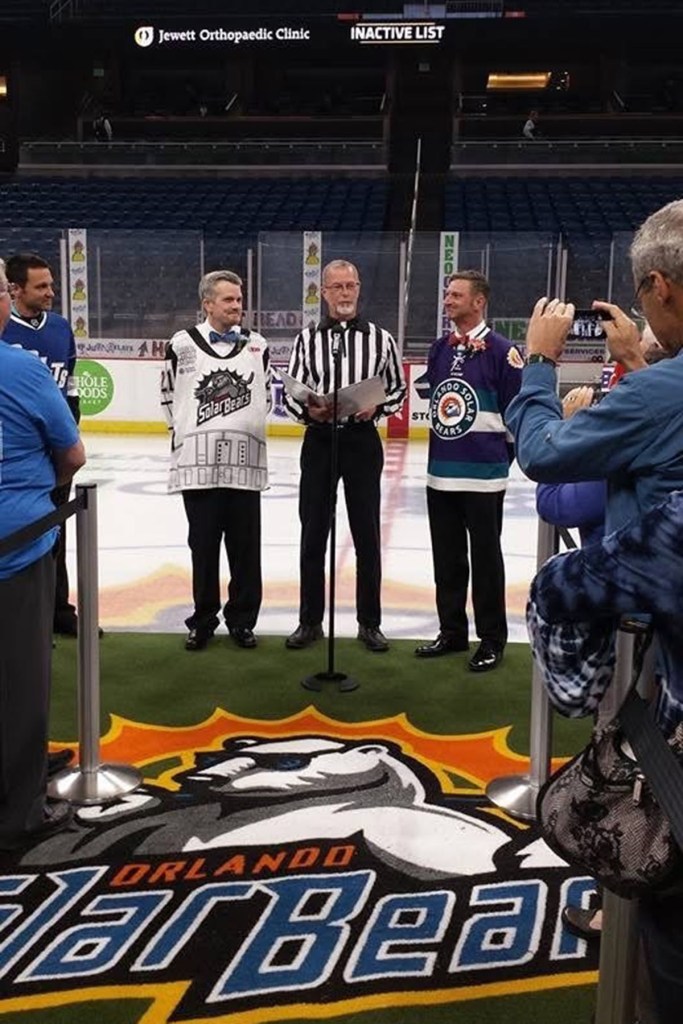 Two men stand on an ice hockey rink during a wedding ceremony, joined by an officiant wearing a referee’s jersey, with spectators nearby and a hockey team logo visible on the ice.