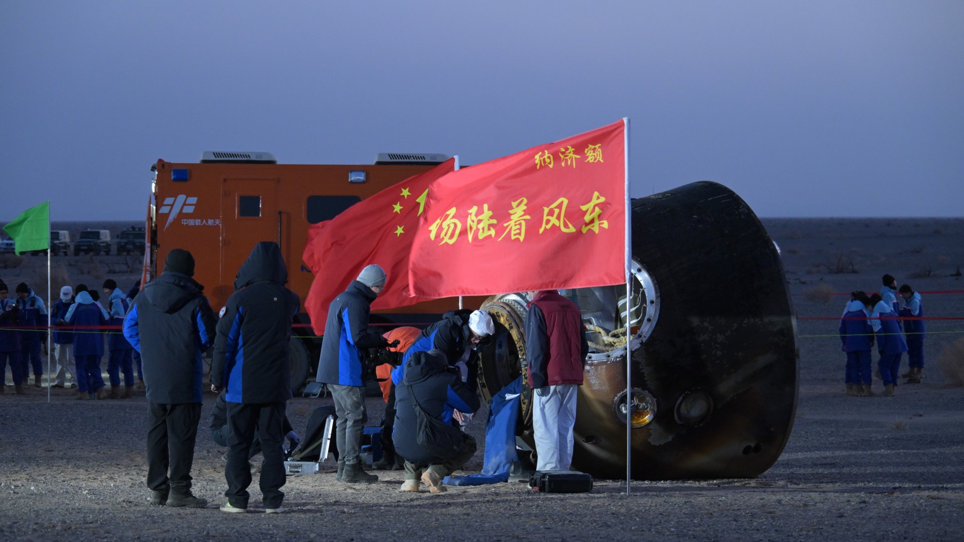 people in heavy coats inspect a charred cone-shaped spacecraft on the floor of the desert