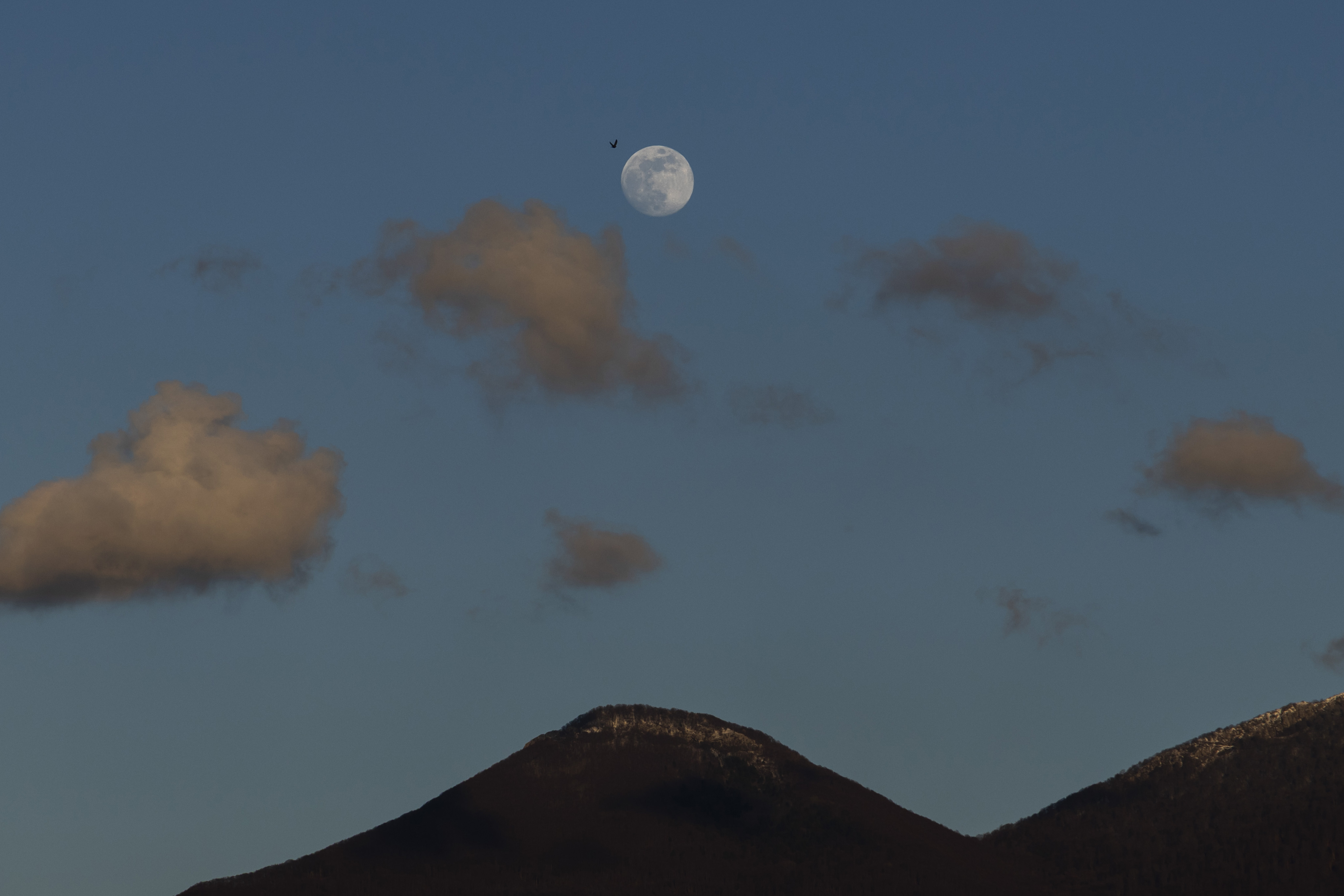 A full moon is pictured above the dark profile of a mountain in the blue evening sky, with small clouds visible below.