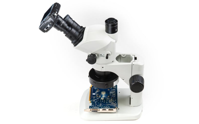 A digital camera is attached to a stereomicroscope, which is focused on a blue electronic circuit board placed on the microscope's stage. The background is white.