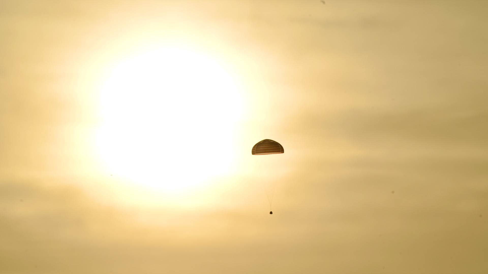 a cone-shaped spacecraft descends under a parachute in front of a setting sun