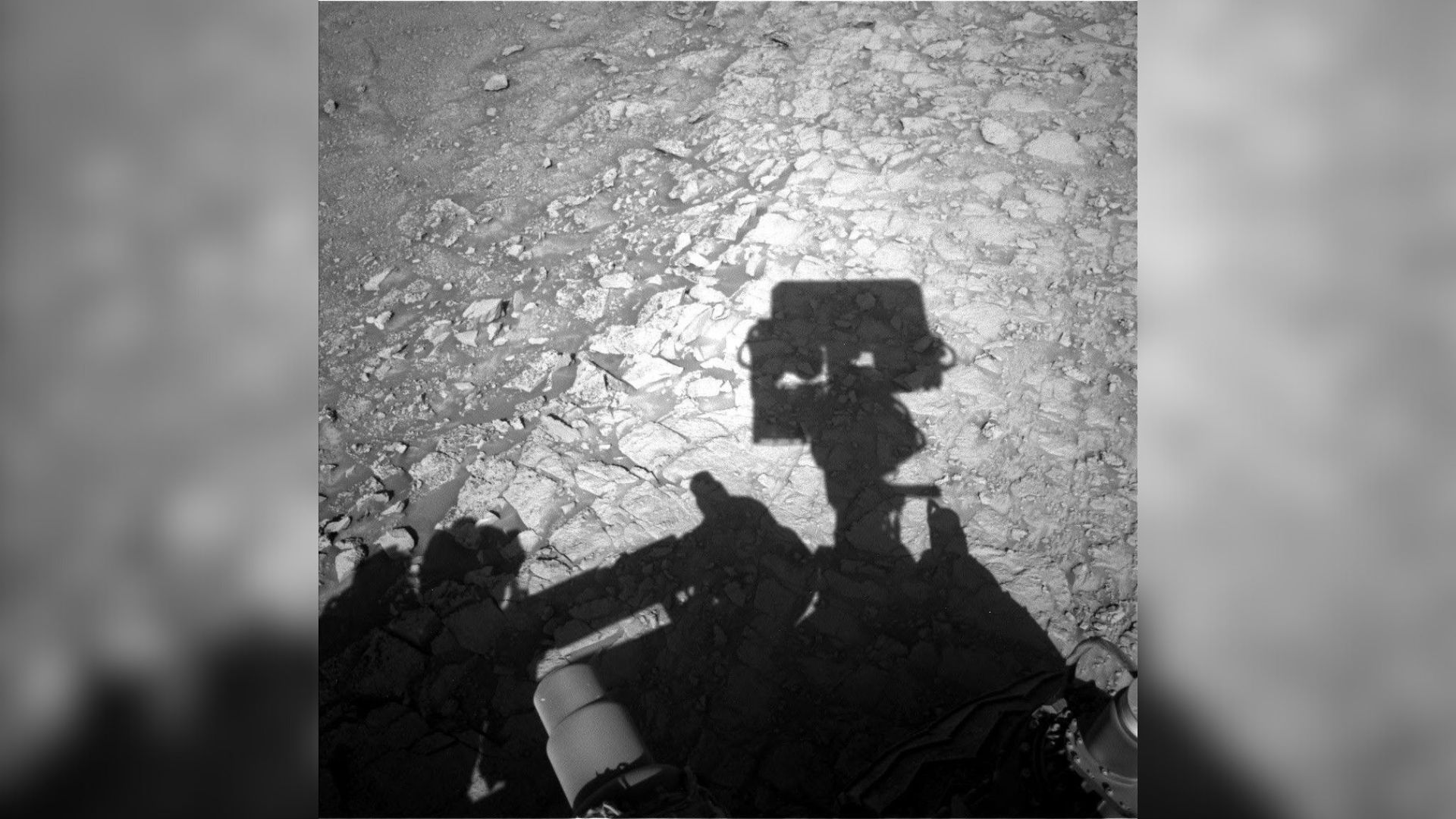 a shadow of a rectangular camera-like appendage on the end of a robotic arm, overlaid on a rocky terrain crisscrossed with tiny ridges