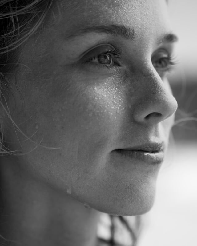 A close-up, black and white photo of a woman's face with water droplets on her skin. She is gazing thoughtfully into the distance, showing subtle freckles and a slight smile, with natural light highlighting her features.