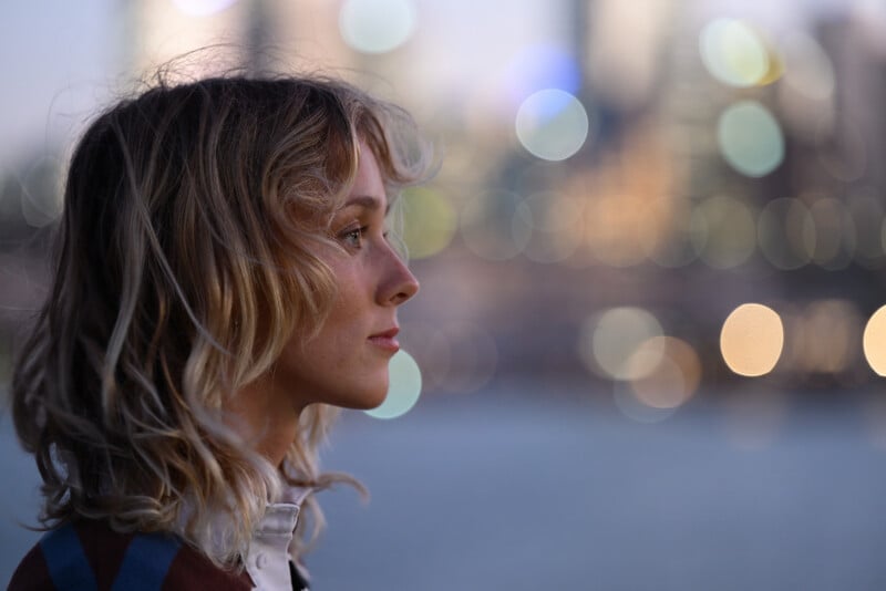 A woman with wavy blonde hair gazes thoughtfully into the distance. The background is blurred with colorful bokeh lights, suggesting an urban setting at dusk or evening.
