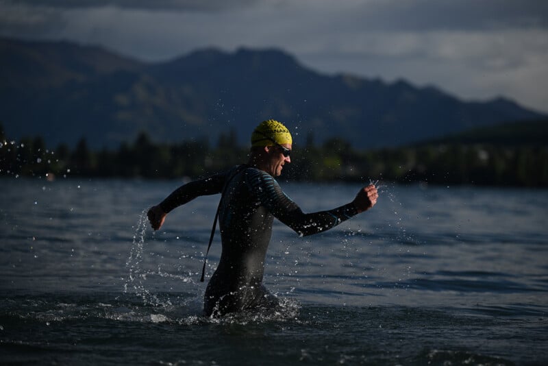 A swimmer wearing a wetsuit, yellow swim cap, and goggles emerges from a lake, splashing water, with mountains and trees visible in the background under a cloudy sky.