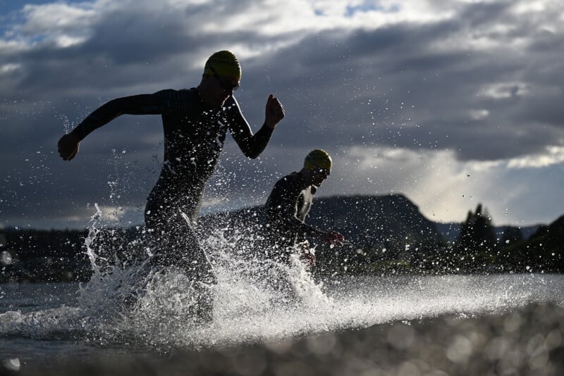 Two athletes in wetsuits and yellow swim caps run through splashing water during a race, with dramatic clouds and a mountainous landscape in the background.