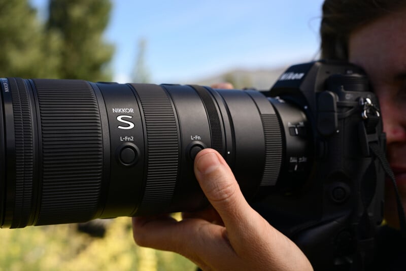 A close-up of a person holding a Nikon camera with a large NIKKOR S telephoto lens, focusing outdoors with blurred greenery in the background.